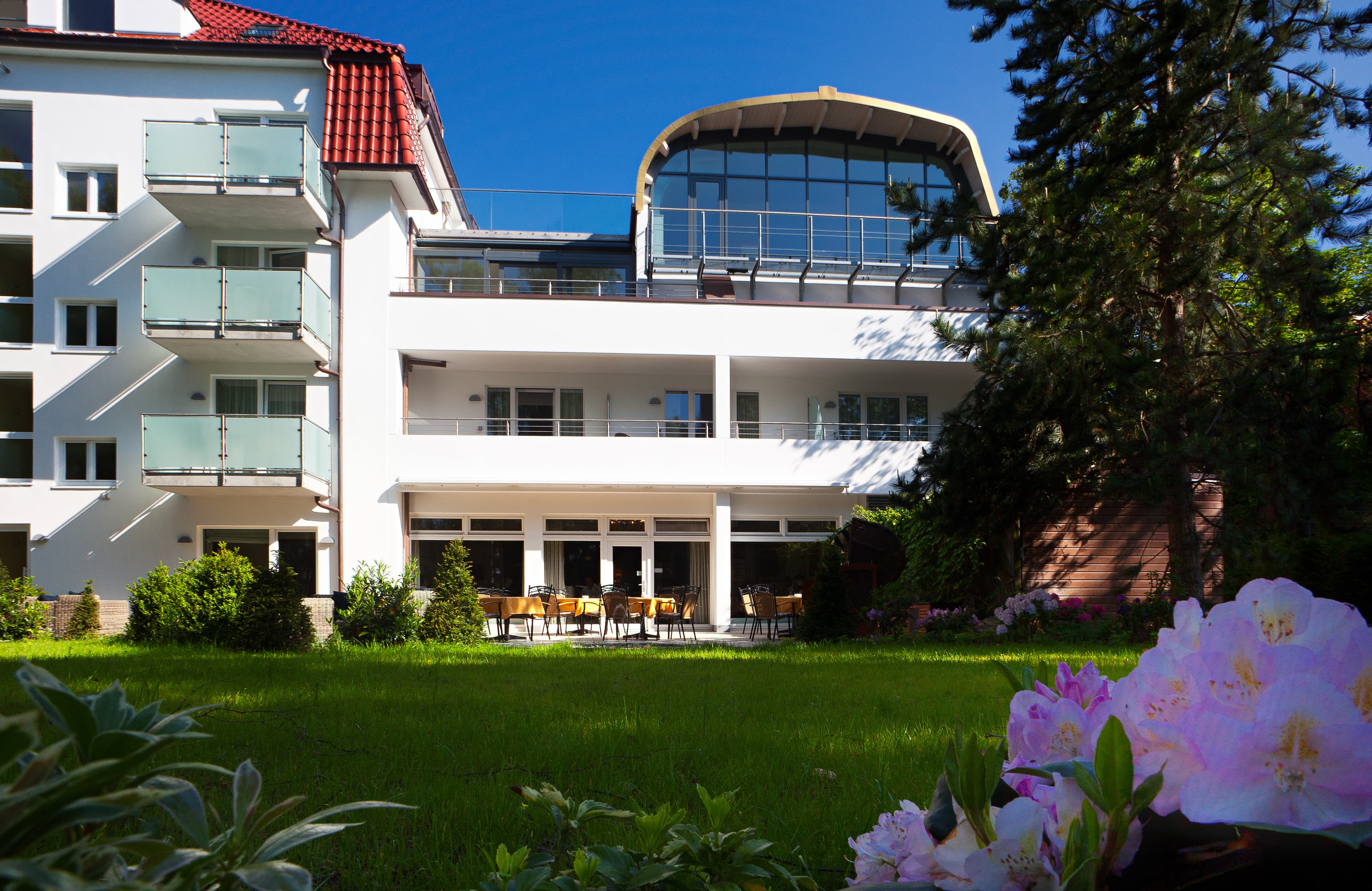 Weißes Gebäude mit Balkonen und Glasaufbau, davor Rasen und Rhododendron